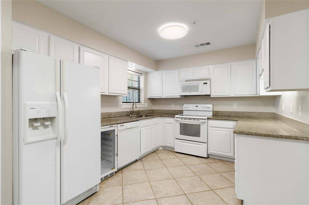 White kitchen featuring white appliances, including a refrigerator with water dispenser, dishwasher, built-in microwave, electric range and granite countertops. The kitchen also has tile flooring and a window for natural light.