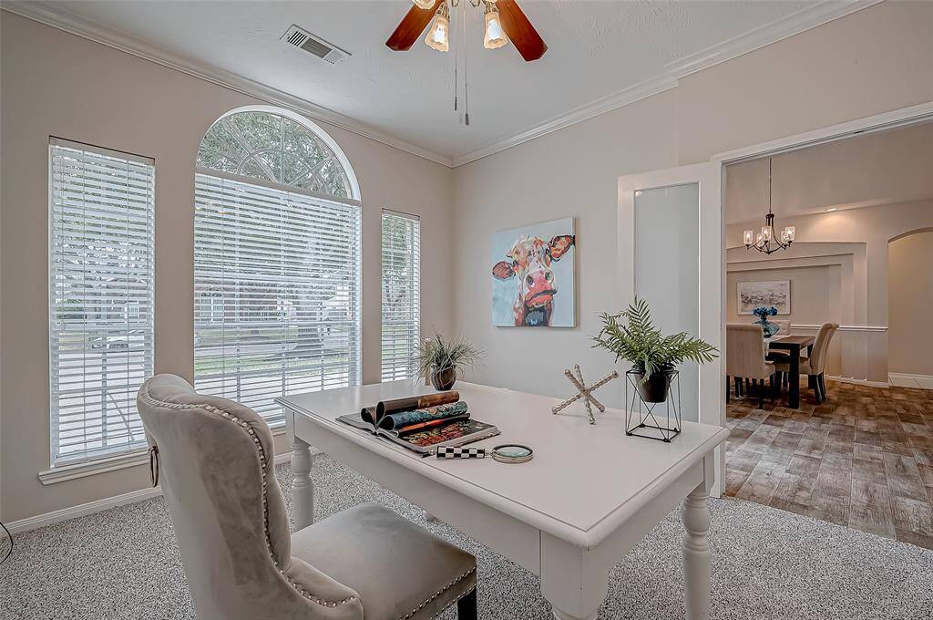 This spacious home office features abundant natural light streaming through a wall of windows with blinds, a ceiling fan for added comfort, plush carpet, and elegant French doors, creating a bright and inviting workspace.