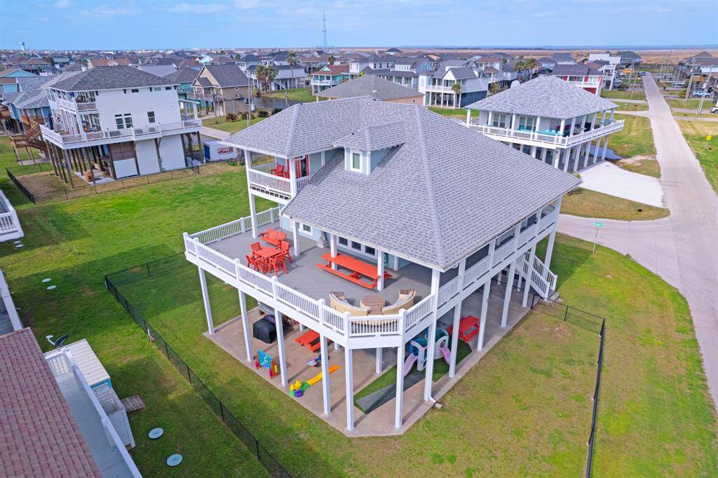This elevated home features a spacious wraparound deck, ideal for outdoor living. The property is located in a deed restricted neighborhood with similar raised homes, offering views of the surrounding area. The yard is fenced, and the house has ample space underneath for storage or recreational use.