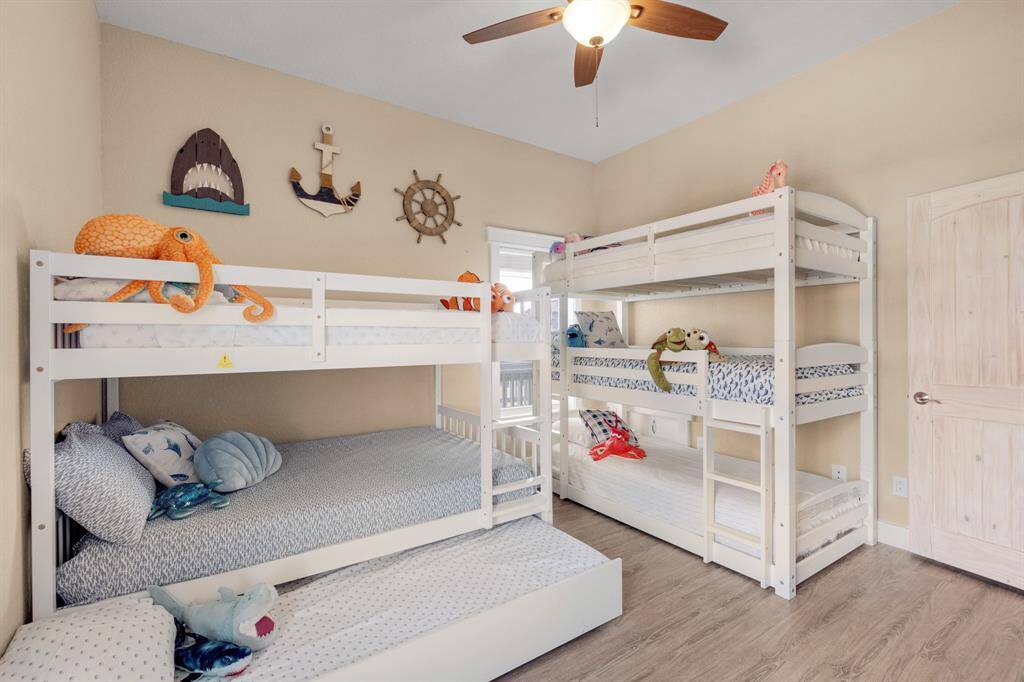 This is a cozy, nautical-themed bedroom featuring two sets of white bunk beds, perfect for kids. The room has light wood flooring and playful marine decor on the walls, creating a fun and inviting atmosphere. A ceiling fan provides comfort and the natural light brightens the space.