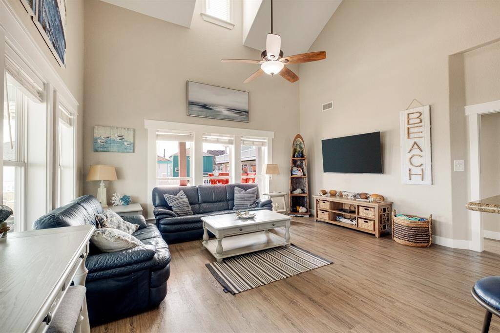 This spacious living room features high ceilings and abundant natural light. It has a cozy coastal decor. The room opens to a bright outdoor area, enhancing its airy feel.