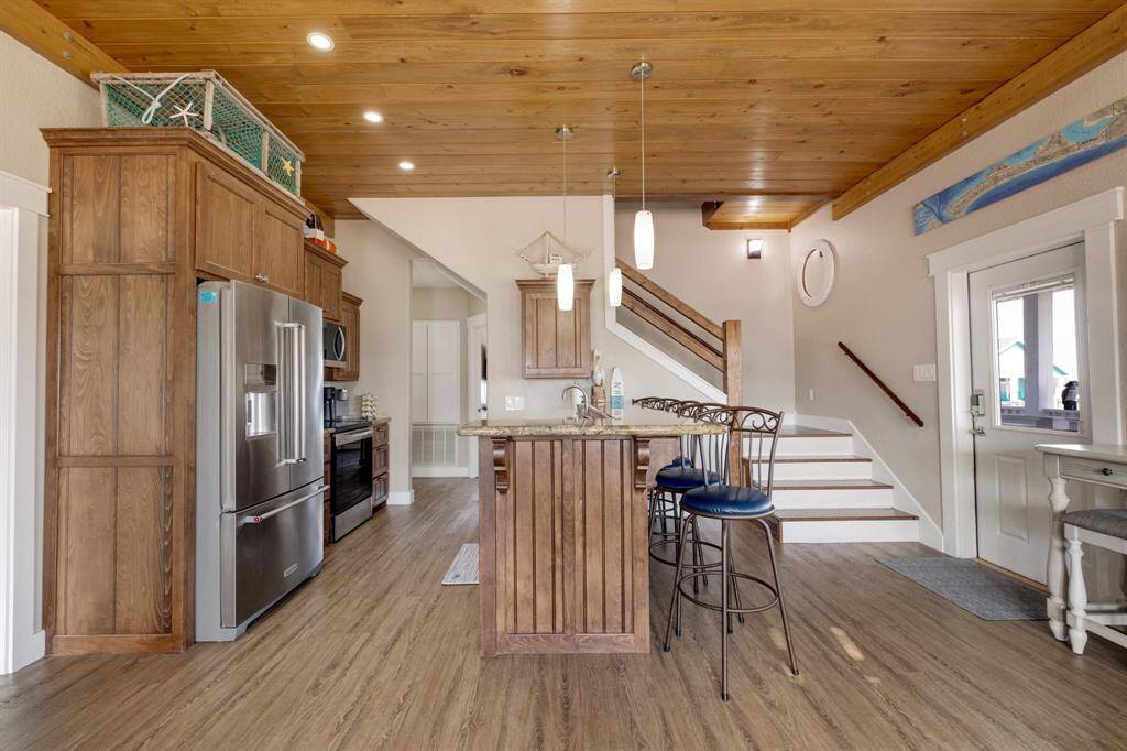 This cozy kitchen features wooden cabinetry, stainless steel appliances, and a center island with bar seating. It has a warm wooden ceiling, pendant lighting, and an open staircase leading to the upper level. The space is bright and inviting, with a door leading to an outdoor area.