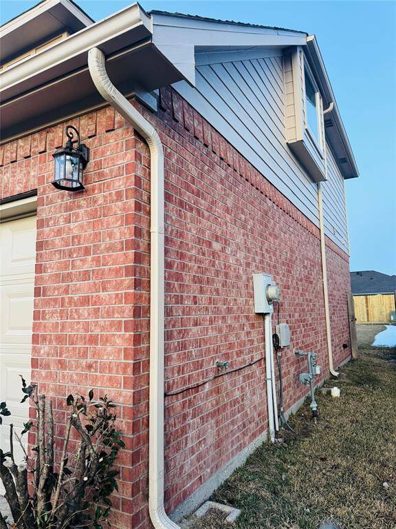 This is a photo of a two-story home with a red brick exterior and beige siding on the upper level. It features a side-mounted light fixture, a white gutter system, and visible utility connections. The yard is partially visible with some grass and a shrub.