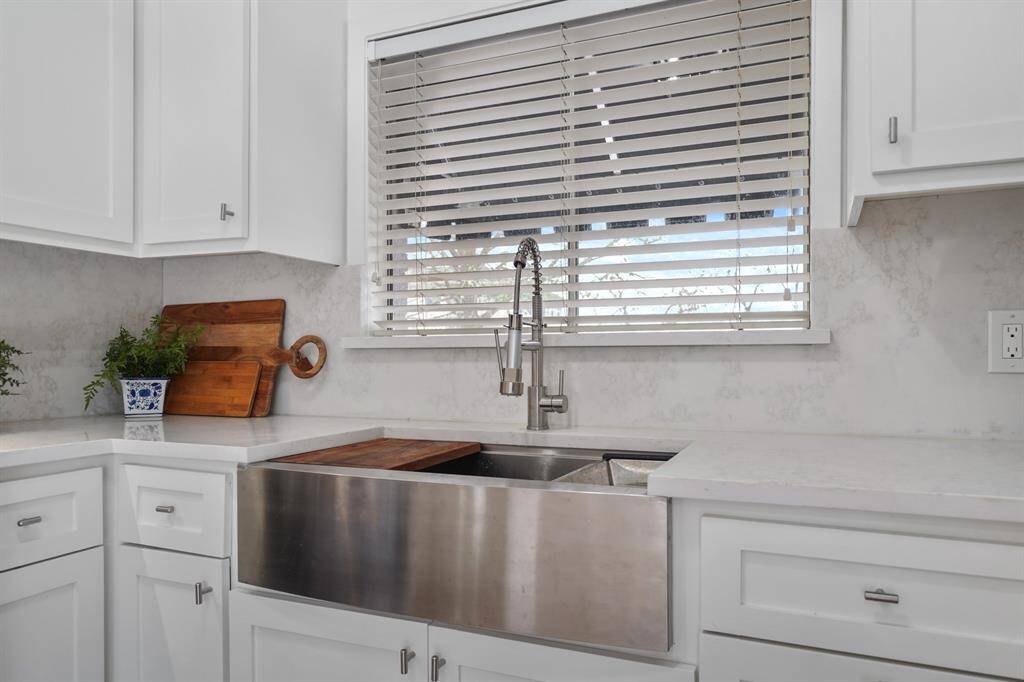 An over-sized stainless steel farm sink sits just below a window with blinds overlooking the front of the home.