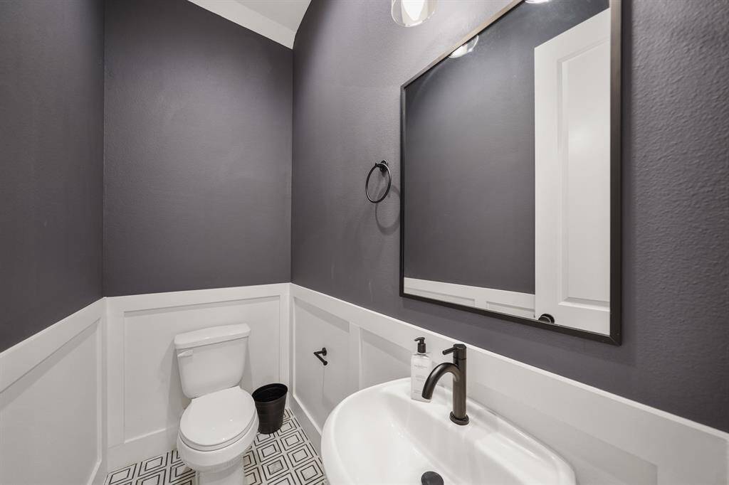 Convenient for guests, the first floor half bath includes a pedestal sink with black plumbing fixtures accompanied by a framed mirror with wall mounted lighting above, wainscoted walls & decorative tile flooring.