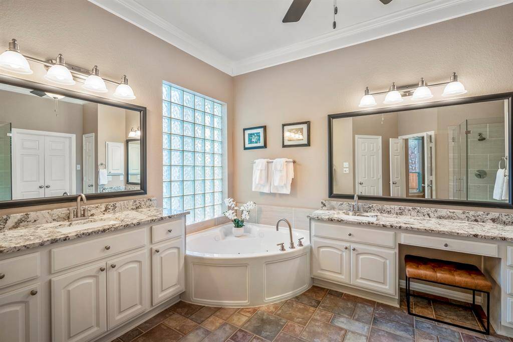 The primary bath offers a luxurious retreat with tile flooring, separate vanities topped with granite counters, and framed mirrors accented by modern light fixtures. A corner jetted tub sits beneath a large window, creating a relaxing oasis. The right-side vanity includes a dedicated seating area, while a linen closet provides extra storage. A frameless glass-enclosed shower adds a sleek, modern touch to this beautifully designed space.