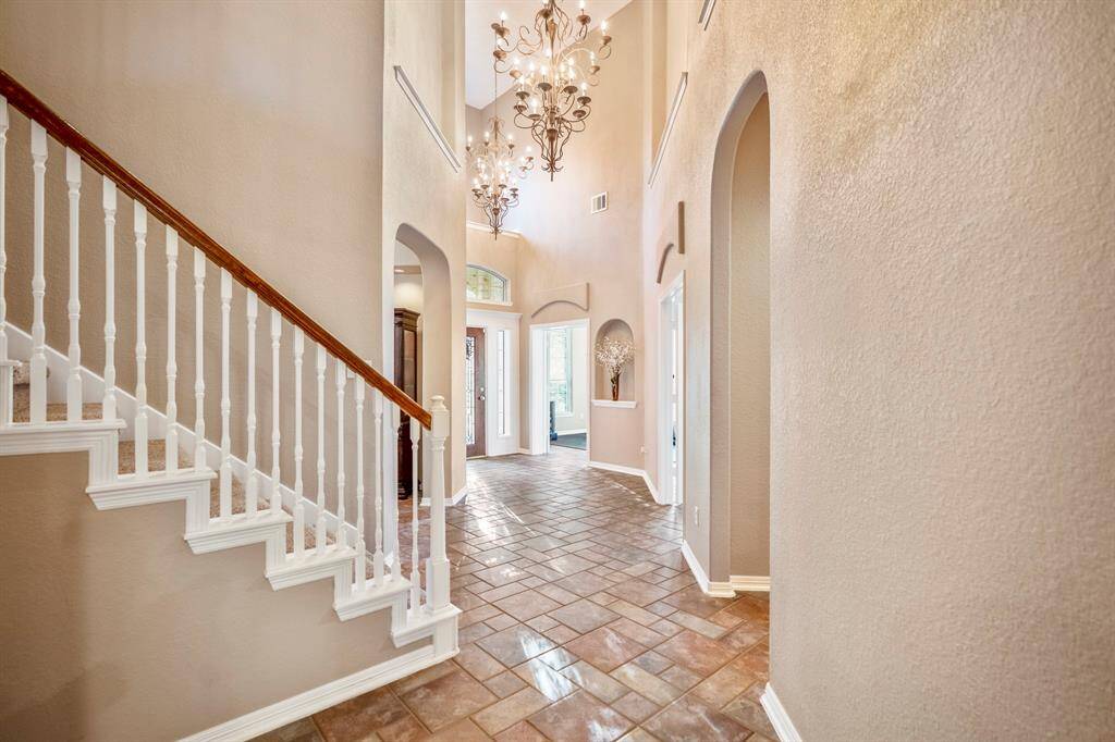 As you move further into the foyer looking back at the front door, the staircase to your left draws the eye, with the formal dining entry just beyond. To your right, a hallway leads to the downstairs half bath and the primary suite. Large windows above and beside the front door flood the space with natural light, highlighting the two striking chandeliers that add drama and elegance to the entry. Through the French doors, glimpses of the formal living and office areas can be seen.