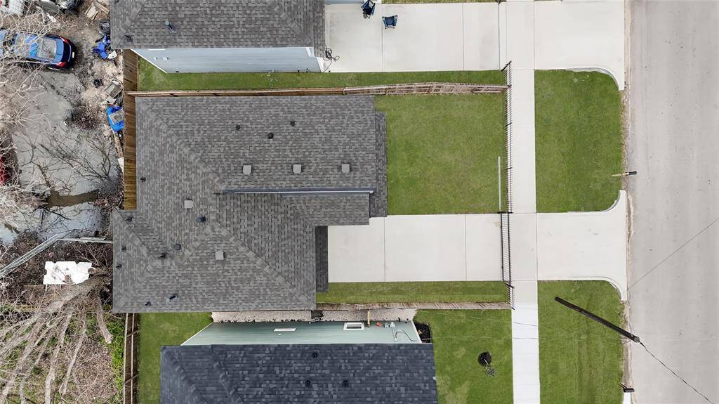 This aerial photo shows a house with a dark shingled roof, a driveway, and a small fenced yard. The property is adjacent to a street with sidewalks and has neighboring homes on both sides.