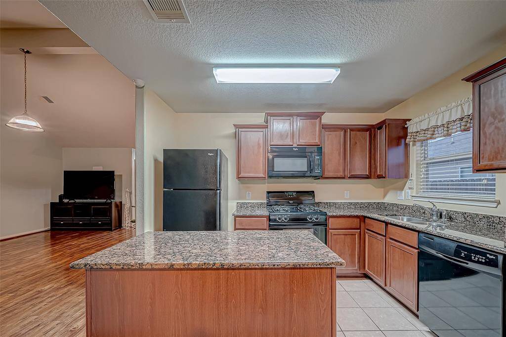 This photo showcases a modern kitchen with granite countertops, dark wood cabinetry, and black appliances. There's an island in the center, and a large window adds natural light. The open layout connects to a living area.
