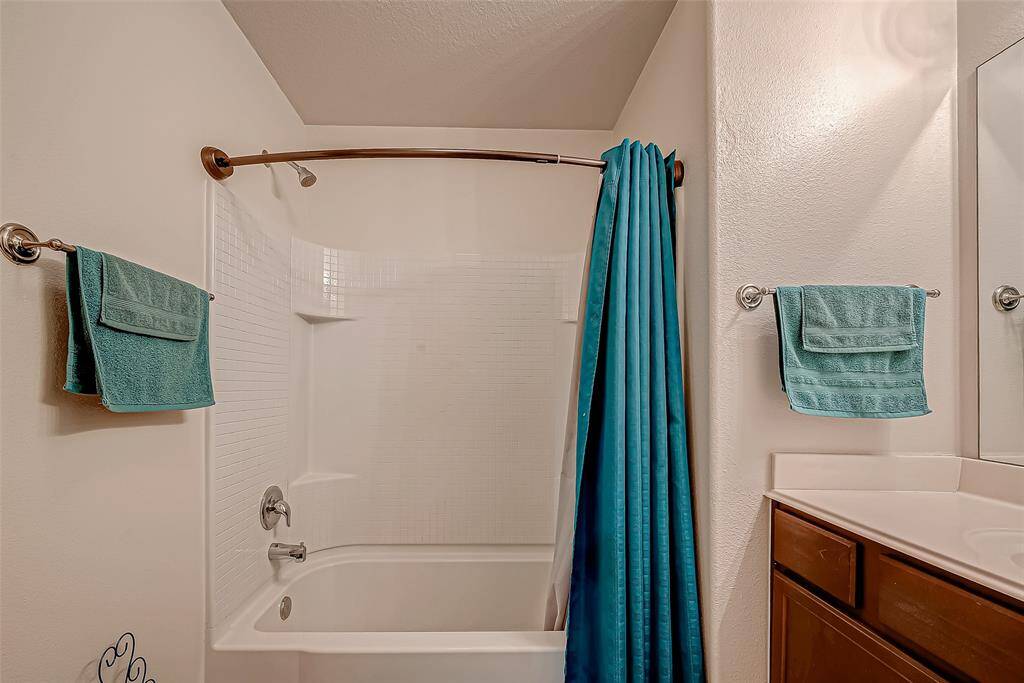 This photo shows a clean, well-lit bathroom featuring a bathtub with a curved shower rod, white tile surround, and teal towels and curtain for a pop of color. The bathroom also includes a vanity with ample counter space.