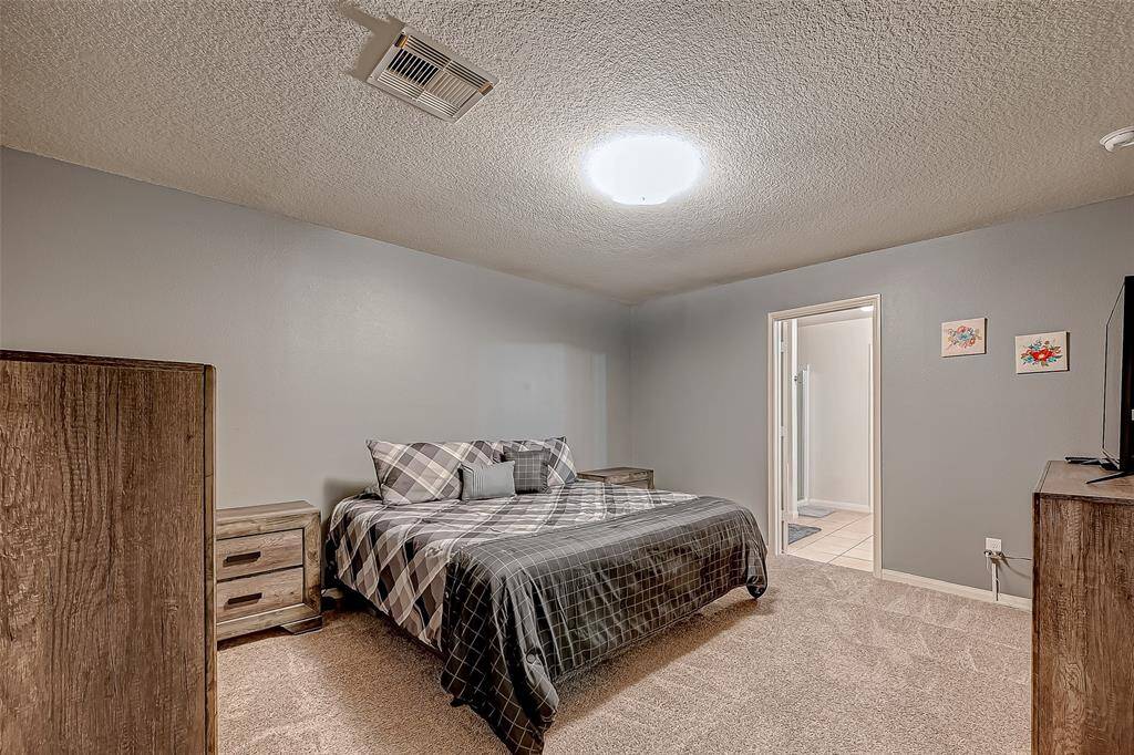 This bedroom features a cozy setup with neutral walls and carpet flooring. It includes a comfortable bed with plaid bedding, wooden furniture, and an attached bathroom. The room is well-lit, creating a warm and inviting atmosphere.