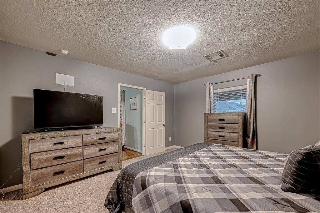 Cozy bedroom with neutral gray walls and carpeted flooring. Includes a large bed with plaid bedding, a wooden dresser with a mounted TV, a window with curtains, and a ceiling light. The room offers a simple, comfortable ambiance.