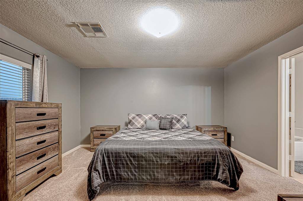 Cozy bedroom with neutral gray walls and carpeted flooring. Features a comfortable bed with plaid bedding, matching wooden nightstands, and a dresser. A window allows natural light, and there's an adjacent bathroom entrance.