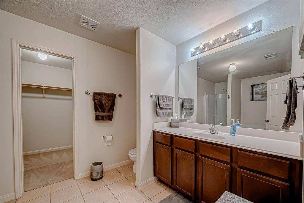 This bathroom features a large vanity with dual sinks and ample storage. It has a spacious mirror with bright lighting fixtures. The room includes a walk-in closet and a separate area for the shower and toilet, all in a neutral color palette.