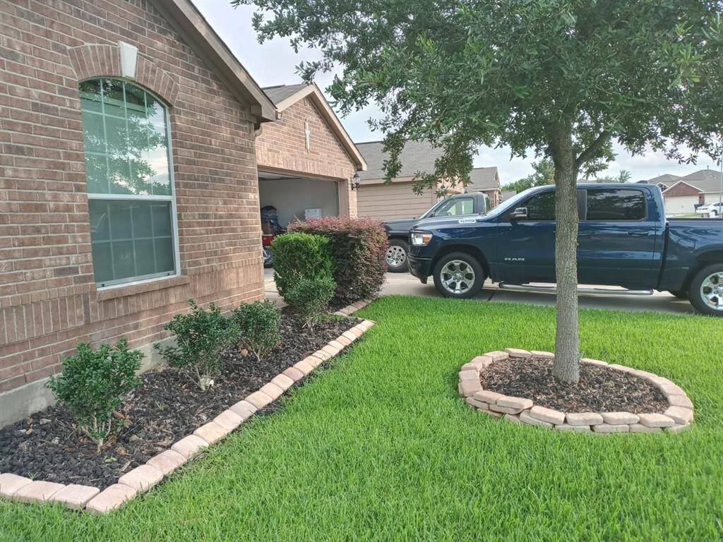 This photo is a spring shot that shows a well-maintained front yard featuring lush green grass and landscaped flower beds with shrubs. A tree is planted in a mulched area bordered with stones.