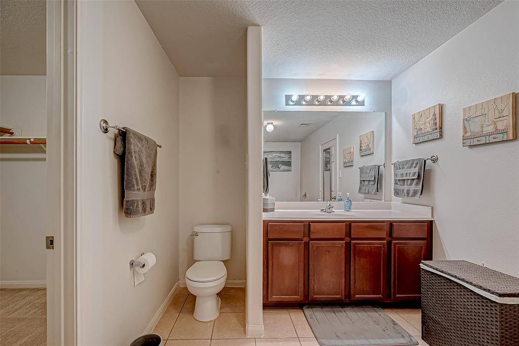 This bathroom features a double-sink vanity with wooden cabinets, a large mirror with overhead lighting, and neutral tile flooring. The space includes a separate toilet area and is accented with wall art and towel racks.