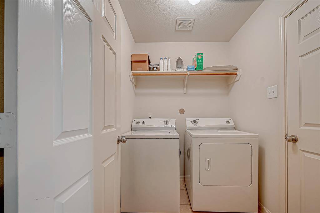 This photo shows a compact laundry room featuring a washer and dryer side by side. There is a shelf above for storage, holding various laundry supplies. The space is well-lit and clean, with neutral walls and two doors.