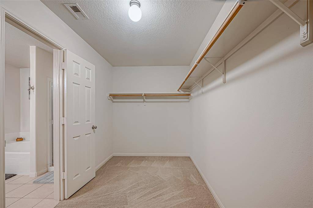 This photo shows a spacious walk-in closet with carpeted flooring, ample hanging space on wooden rods, and an overhead light fixture. It adjoins a tiled bathroom.