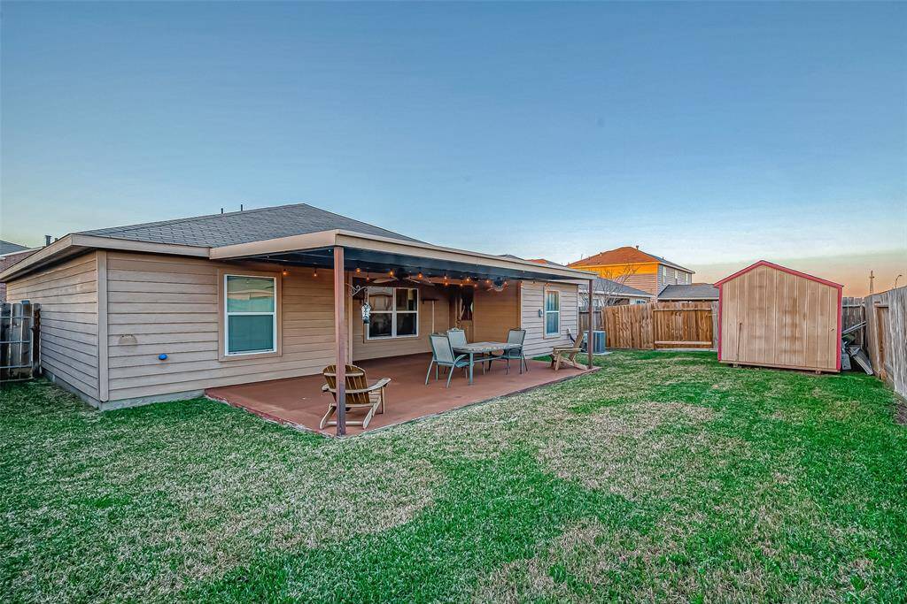 This backyard features a covered patio with seating, a spacious lawn, and a wooden storage shed, all enclosed by a wooden fence, offering a cozy outdoor space for relaxation and storage.