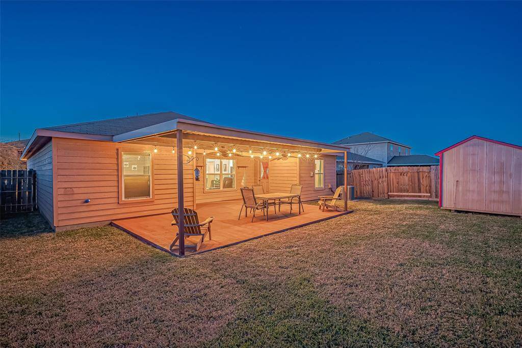 This photo shows a cozy backyard with a covered patio, adorned with string lights, perfect for outdoor relaxation. The lawn is spacious, and there's a small wooden shed, offering additional storage or workspace options.