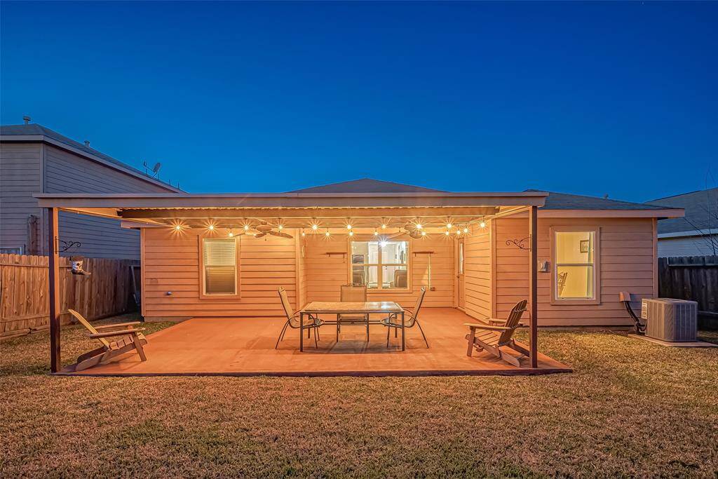 This photo showcases a cozy backyard patio with a covered pergola adorned with string lights, perfect for evening gatherings. The space includes seating and a table, set on a concrete pad, with a grassy area surrounding it, creating an inviting outdoor retreat.