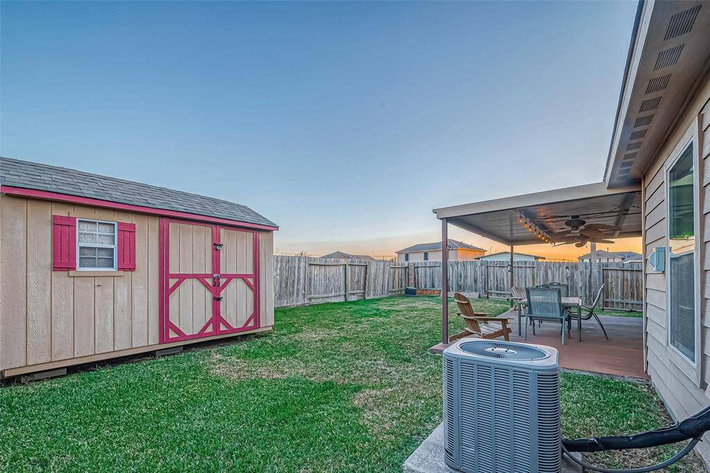 This backyard features a spacious green lawn, a large storage shed with red accents, and a covered patio area ideal for outdoor dining and relaxation. The space is enclosed by a wooden fence, providing privacy.