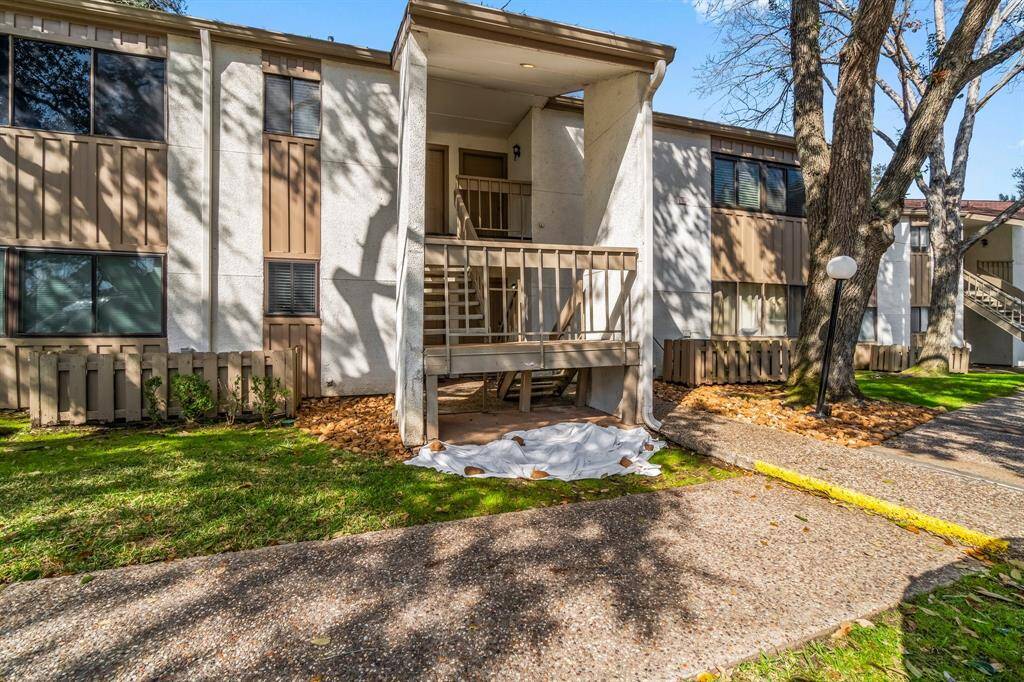 two-story apartment building with a small balcony and a covered entryway. The exterior features a mixed facade of light and dark panels. There's a well-maintained lawn with mature trees, and a paved walkway leading to the entrance.