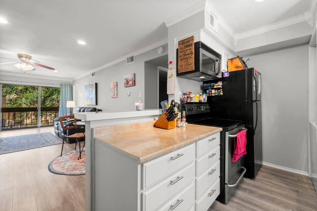 Cozy open-plan kitchen and living room with light wood flooring and soft gray walls. The kitchen features a black appliance suite and white cabinetry, while the living area includes a ceiling fan and large window offering natural light and views of greenery.