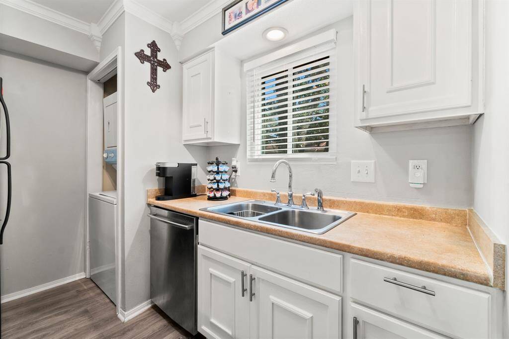 This kitchen features sleek white cabinetry, a double stainless steel sink, and Quartz countertops. It includes a compact dishwasher. A window above the sink provides natural light, and there's a glimpse of a stacked washer and dryer set in an adjacent nook.