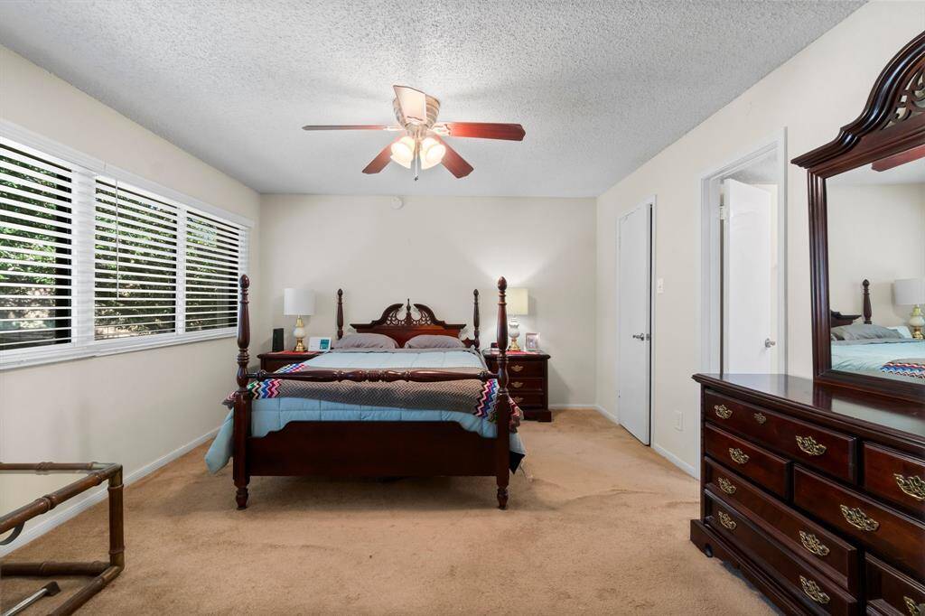 Primary bedroom. The room is well-lit by a large window with blinds, and a ceiling fan offers comfort. Neutral walls and carpet create a warm atmosphere.