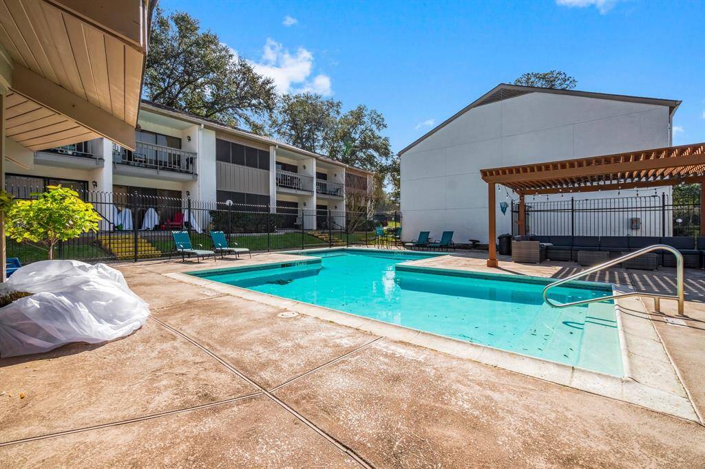 This photo showcases a communal outdoor pool area with a sun deck and lounge chairs, surrounded by a fence. The setting is part of a residential complex, offering a relaxing space for residents with nearby greenery and a pergola for shade.