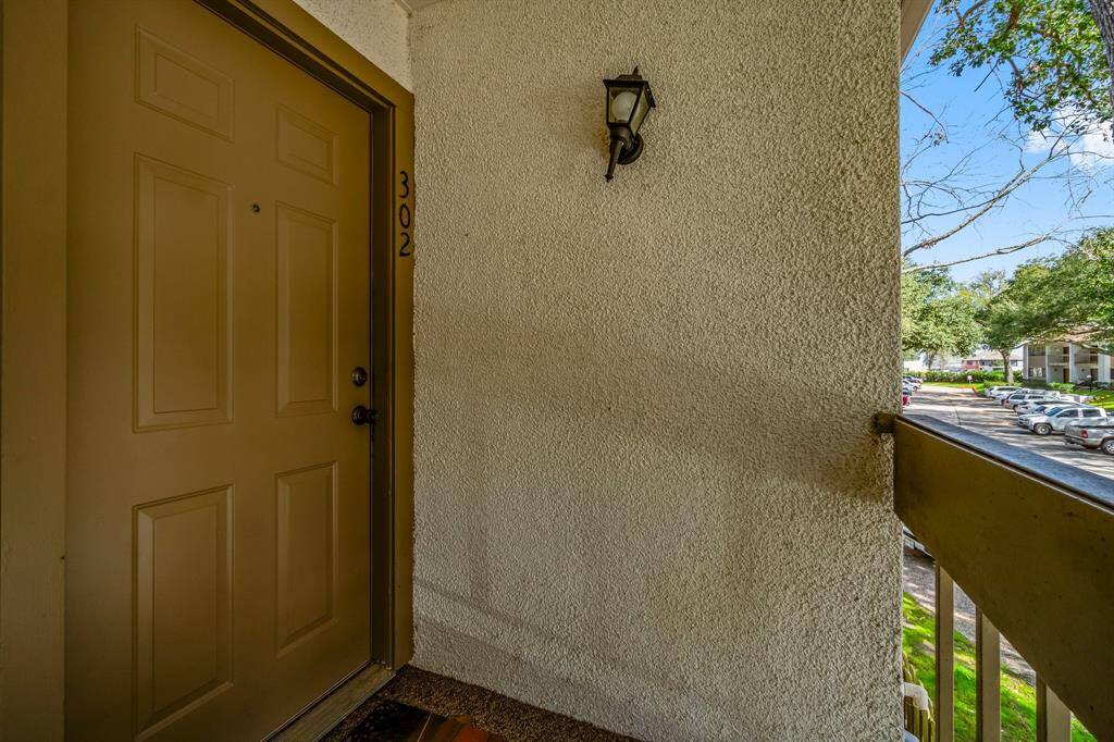 The entryway to a second-floor apartment, featuring a solid front door with a classic outdoor wall lamp. The area overlooks a parking lot with mature trees, providing a pleasant view.
