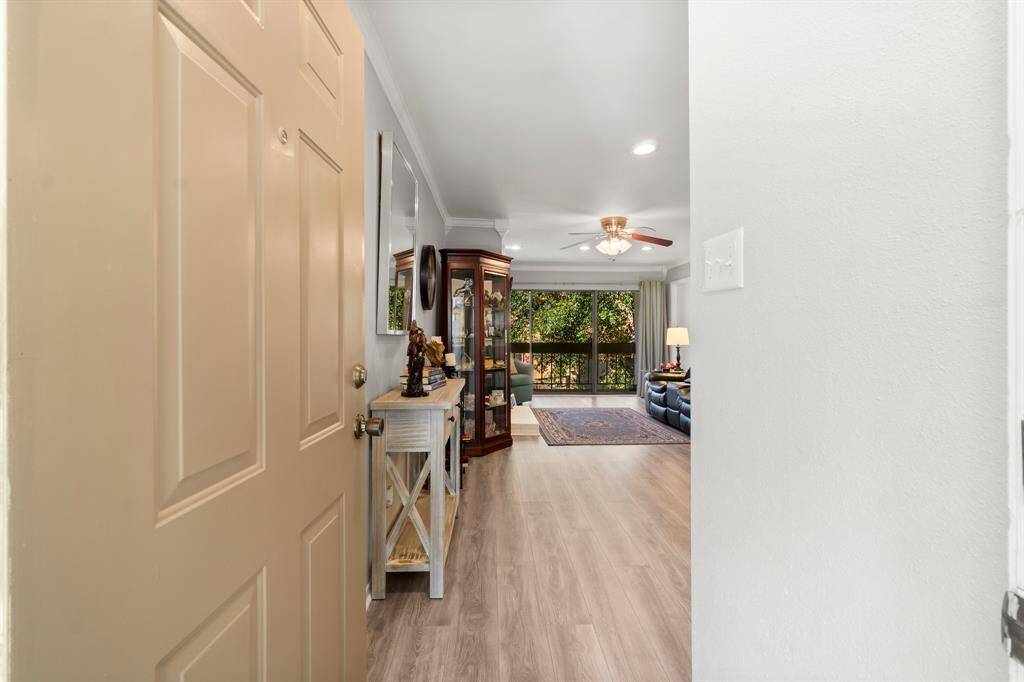 Welcome to a bright, inviting living space featuring hardwood floors and a ceiling fan. The entryway leads to a cozy, well-lit living room with large windows offering a view of greenery, perfect for relaxation.