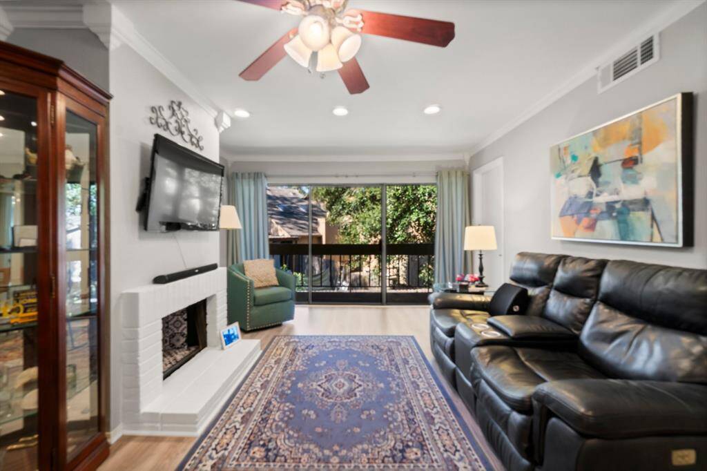 A cozy living room featuring a modern ceiling fan,  The space is accented by a white brick fireplace. Large windows offer natural light and a view of greenery, creating a welcoming atmosphere.