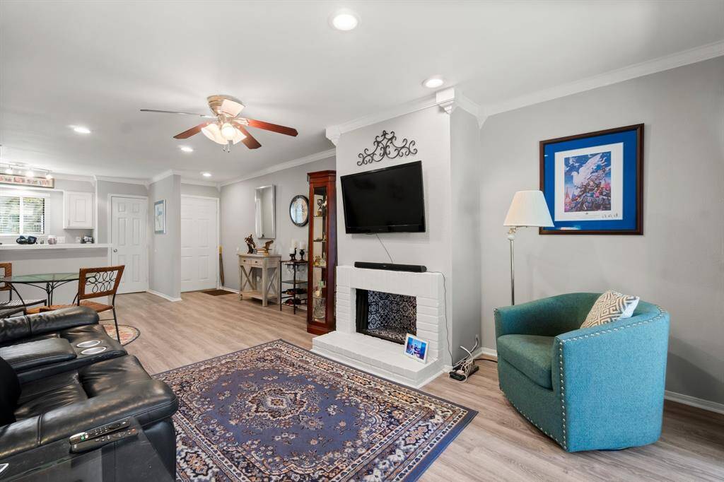 This cozy living room features a modern open layout with a fireplace. The space is bright with recessed lighting and a ceiling fan, and it seamlessly flows into a kitchen area, creating a welcoming environment.