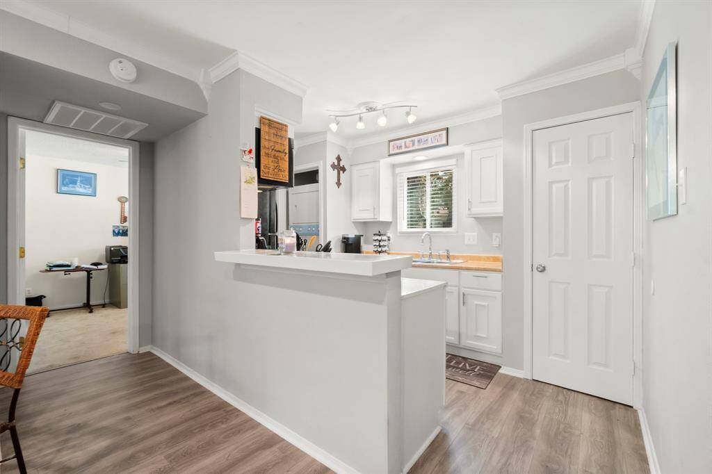 This photo showcases a bright, modern kitchen with white cabinetry and light wood flooring. It features a cozy breakfast bar, ample counter space, and a window above the sink for natural light. Adjacent is a doorway leading to another room, adding convenience and flow to the home's layout.