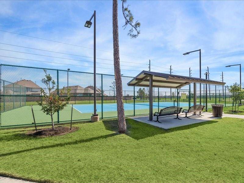 This photo features a well-maintained tennis court with a surrounding fence, adjacent to a covered seating area on a neatly landscaped lawn, ideal for outdoor recreation and relaxation.
