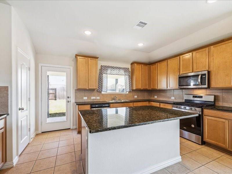 This kitchen features warm wooden cabinetry, a large granite island, stainless steel appliances, and a tile floor. It includes a window over the sink and a door leading outside, providing ample natural light.