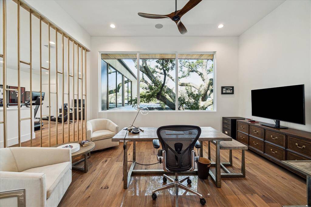 A glimpse into the study, where warm wood flooring, recessed lighting, a modern ceiling fan and Sonos sound speakers create a comfortable workspace. The focal point is a large window with treetop views, bringing in natural light and a tranquil connection to the outdoors.