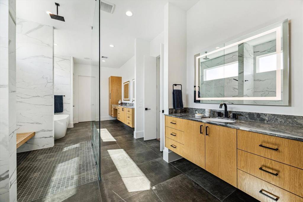 The primary bath exudes luxury, featuring a double-door entry, and matte porcelain flooring leading to thoughtfully designed separate spaces. It boasts two vanities with Quartzite countertops and LED-lit mirrors, two private water closets, and two spacious walk-in closets outfitted with extensive built-ins.