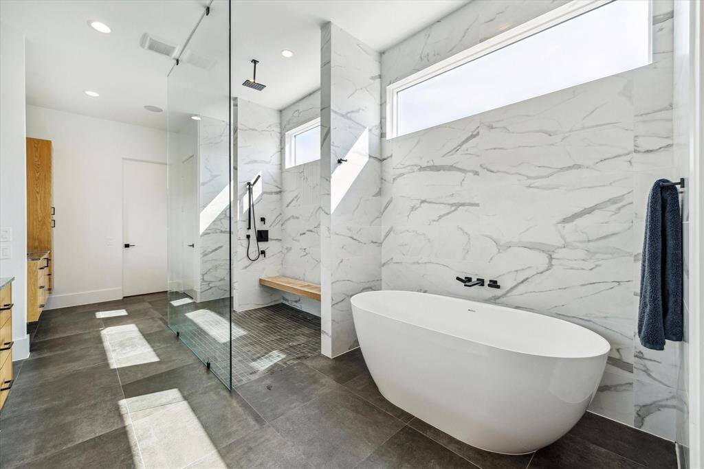 A true spa-like experience, the primary bath showcases a relaxing free-standing soaking tub and an expansive walk-in glass shower with bench seat, elegantly lined with glazed ceramic tile in a matte marble finish.