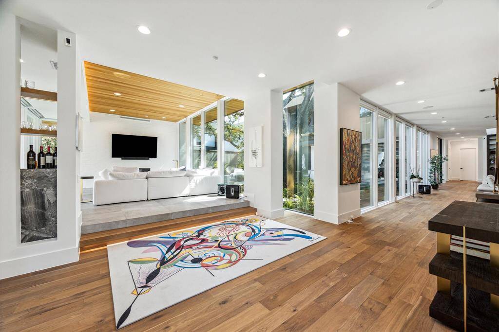 A striking entrance welcomes you with warm wood flooring and expansive glass walls that flood the space with natural light, creating a seamless indoor-outdoor connection. To the left, a lounge area with a wood-clad ceiling offers a cozy yet modern retreat, while the open hallway to the right leads to inviting living areas.