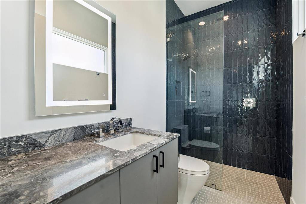 The room's ensuite bath is complete with matte grey ceramic flooring, a walk-in glass shower wrapped in high-gloss ceramic subway tile, a Quartzite-topped storage vanity, and an LED-lit mirror.