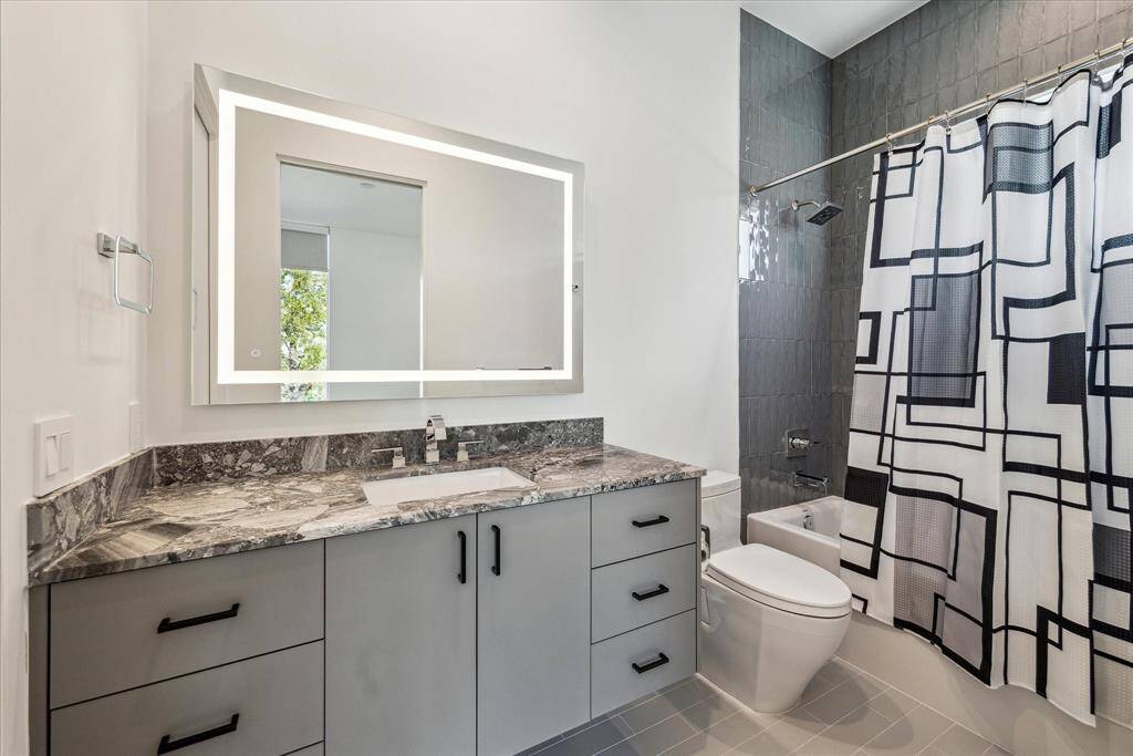 The room's ensuite bath is complete with matte grey ceramic flooring, a tub/shower combination surrounded by high-gloss ceramic subway tile, a Quartzite-topped storage vanity, and an LED-lit mirror.