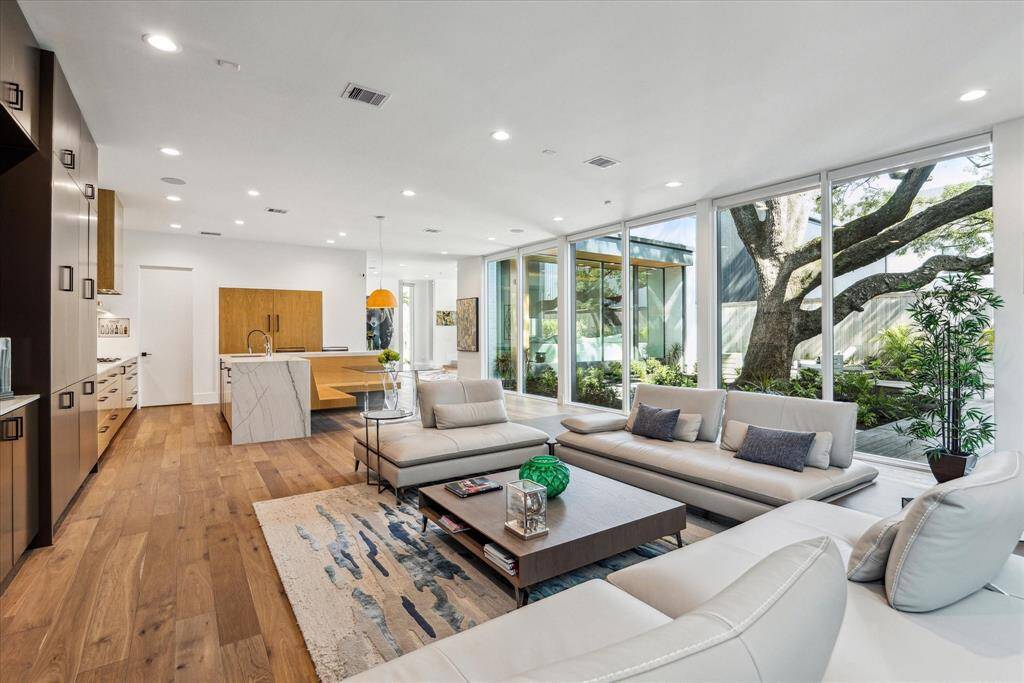The kitchen flows into the living and dining areas and is lined by a wall of glass that overlooks a serene outdoor retreat and a towering oak tree. A wall of built-ins provides efficient and stylish storage solutions, while the airy design fosters a natural connection between the interior and the scenic surroundings.