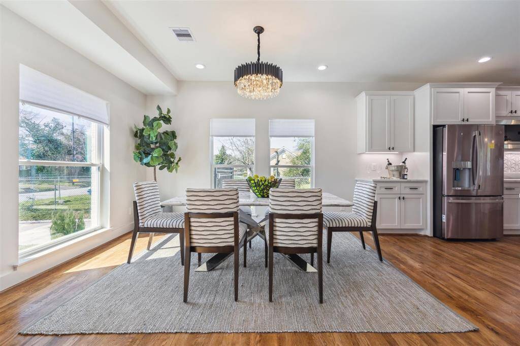 This photo showcases a bright, modern dining area with large windows, a stylish chandelier, and a cozy rug. The dining set features contemporary chairs around a glass table, and the open layout includes a sleek kitchen with stainless steel appliances and white cabinetry.