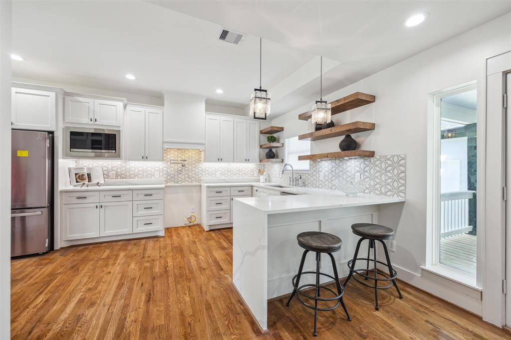 This kitchen features modern white cabinetry, a stylish tile backsplash, and stainless steel appliances. It has a breakfast bar with two stools, pendant lighting, and open shelving for a contemporary touch. The hardwood floors add warmth to the space, and a large window brings in natural light.