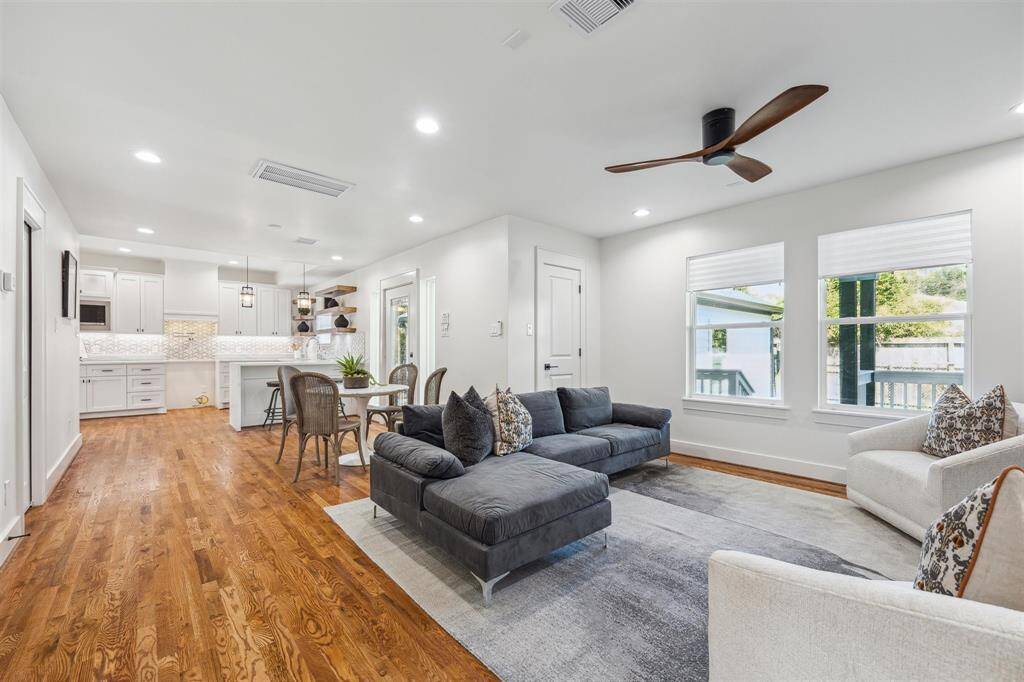 This photo showcases a bright, open-concept living space with hardwood floors, featuring a modern kitchen with white cabinetry and a cozy living area. Large windows provide ample natural light, and the space is tastefully furnished with contemporary decor.