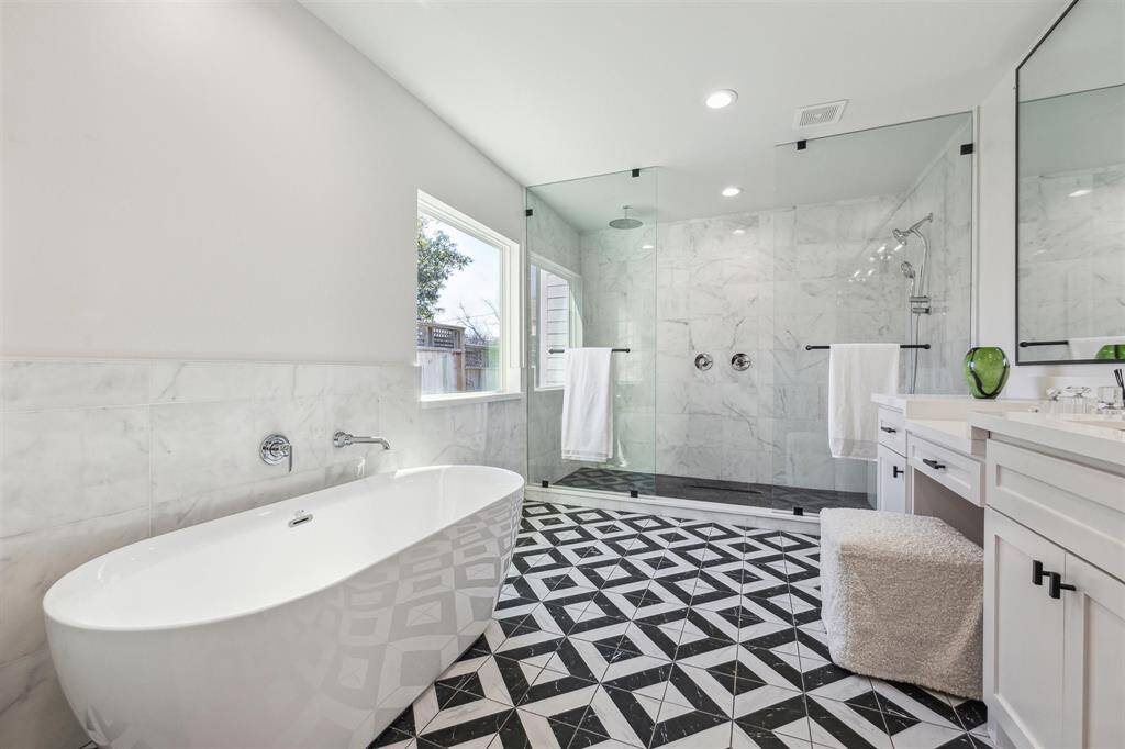 This modern bathroom features a spacious walk-in shower with glass doors, a sleek freestanding bathtub, and stylish black-and-white geometric floor tiles. Ample natural light streams in through a large window, complementing the contemporary white cabinetry and marble accents.