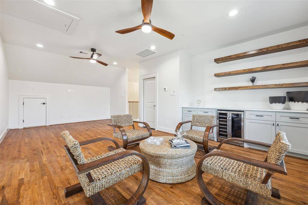 This spacious room features warm wooden flooring and a cozy seating area.  The room includes a modern wet bar with a wine cooler, open shelving, and sleek cabinetry, all under soft recessed lighting and ceiling fans.